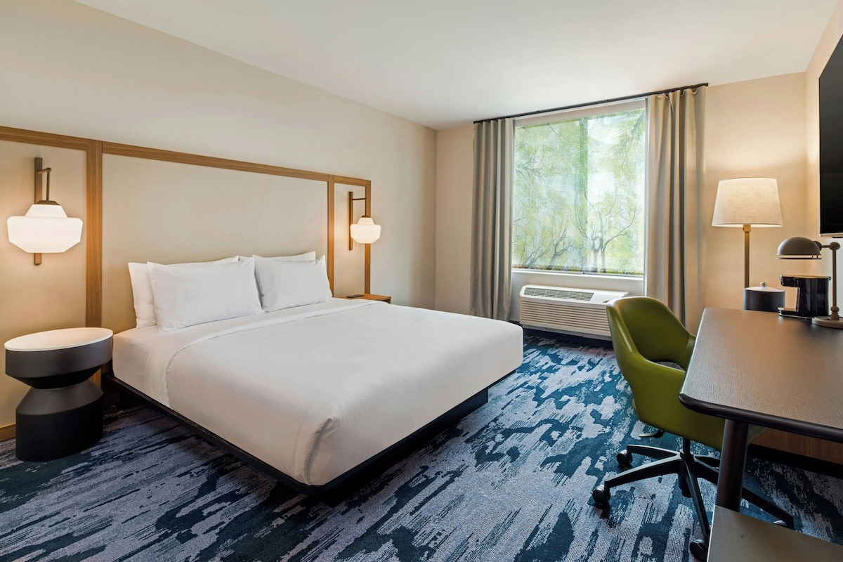 Quarto de hotel com cama, mesas de cabeceira, escrivaninha, cadeira, janela com cortinas e luminária na parede.
