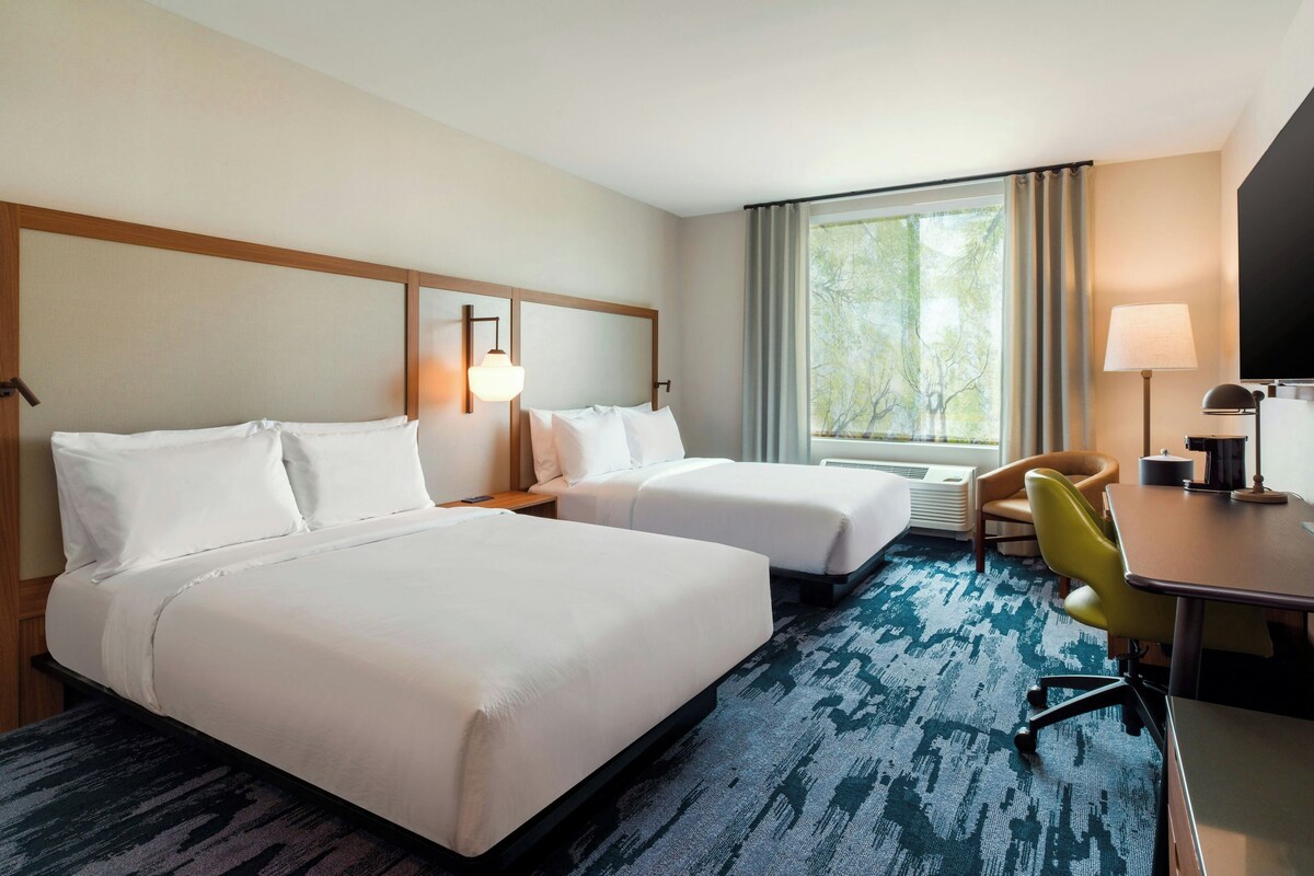 Quarto de hotel com duas camas, uma escrivaninha, uma cadeira, um abajur e uma janela grande com cortinas.