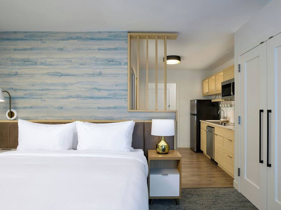 A hotel room with a bed, bedside lamp, and a view of the kitchen.