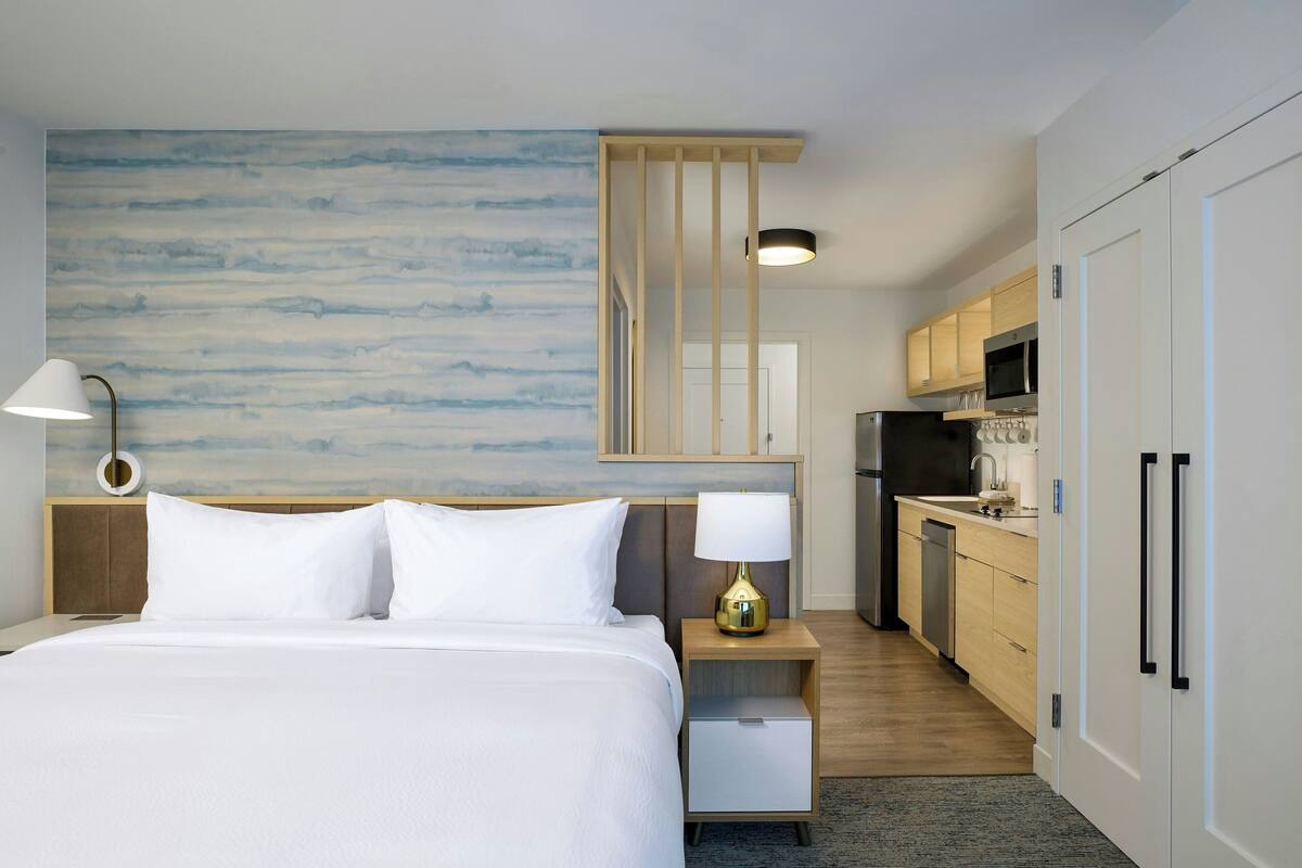 Ein Hotelzimmer mit Bett, Nachttisch mit Lampe, einer kleinen Küchenzeile mit Mikrowelle und einem Holzschrank.