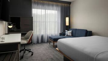 A hotel room with a bed, a desk, a chair, a TV, and a window with curtains.
