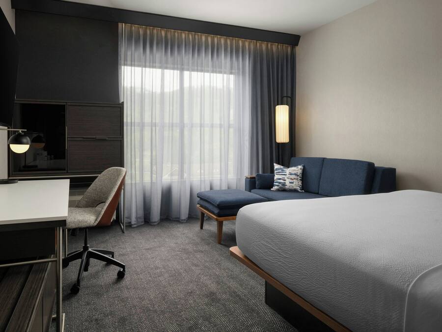 A hotel room with a bed, a desk, a chair, a TV, and a window with curtains.