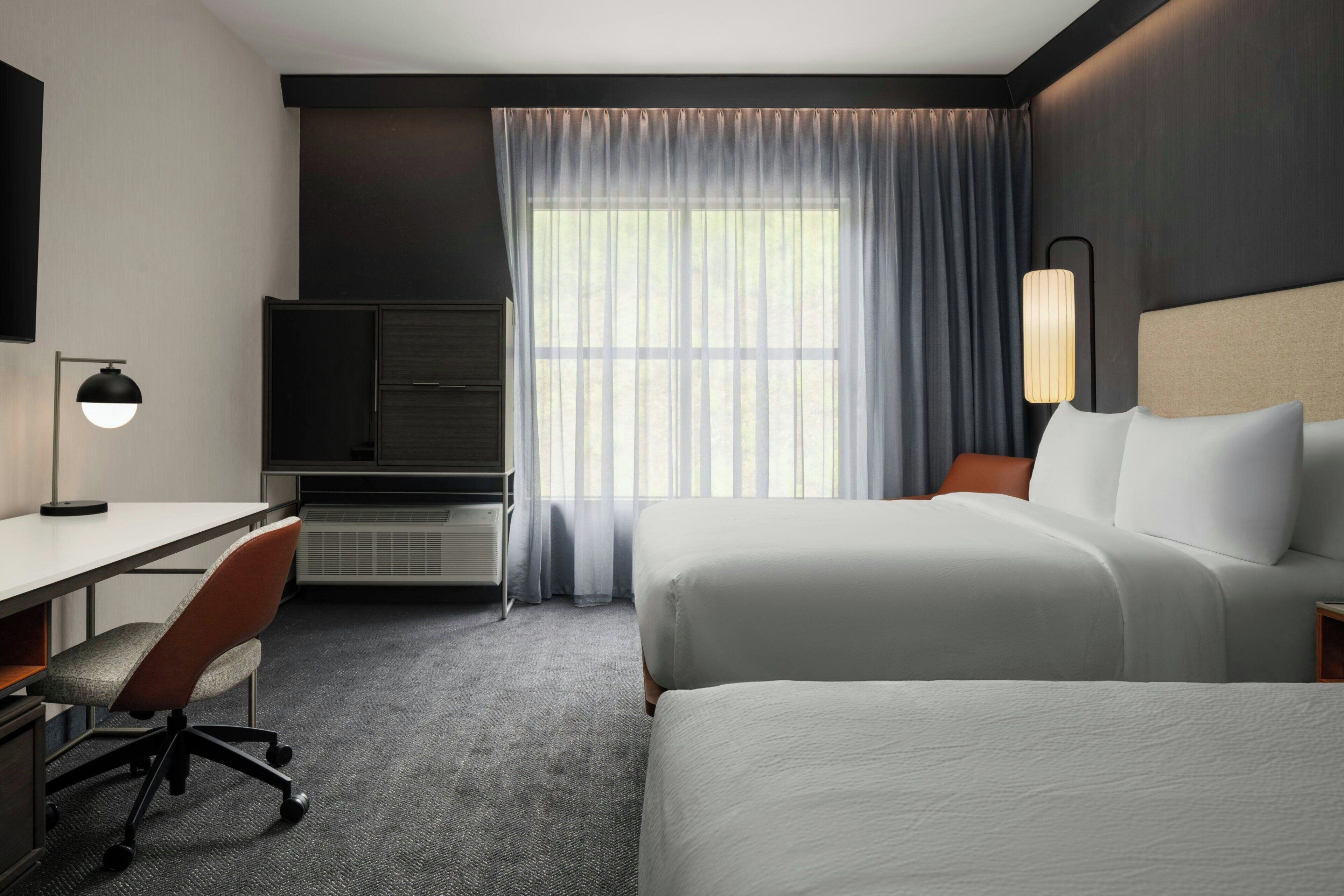A hotel room with a bed, desk, chair, and a large window with curtains.