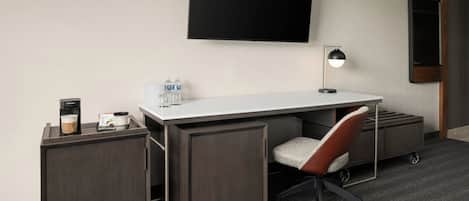 A hotel room with a desk, chair, TV, coffee machine, and water bottles.