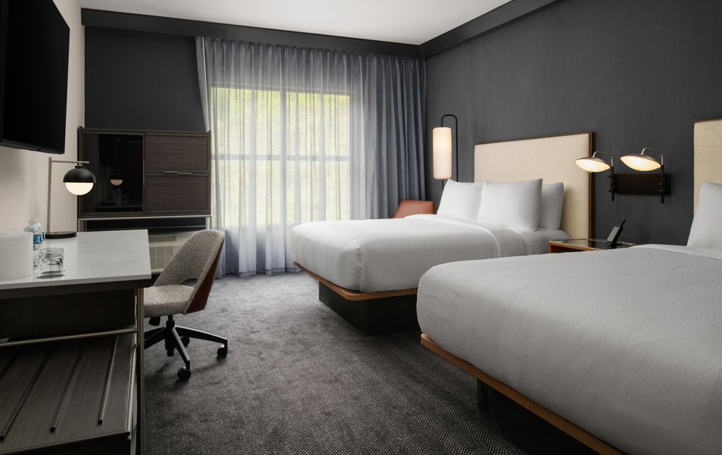 A hotel room with two beds, a desk, a chair, a TV, and a window with curtains.