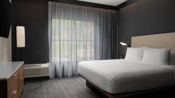 Une chambre d’hôtel avec un lit, un bureau, une chaise, une lampe et une fenêtre avec des rideaux.
