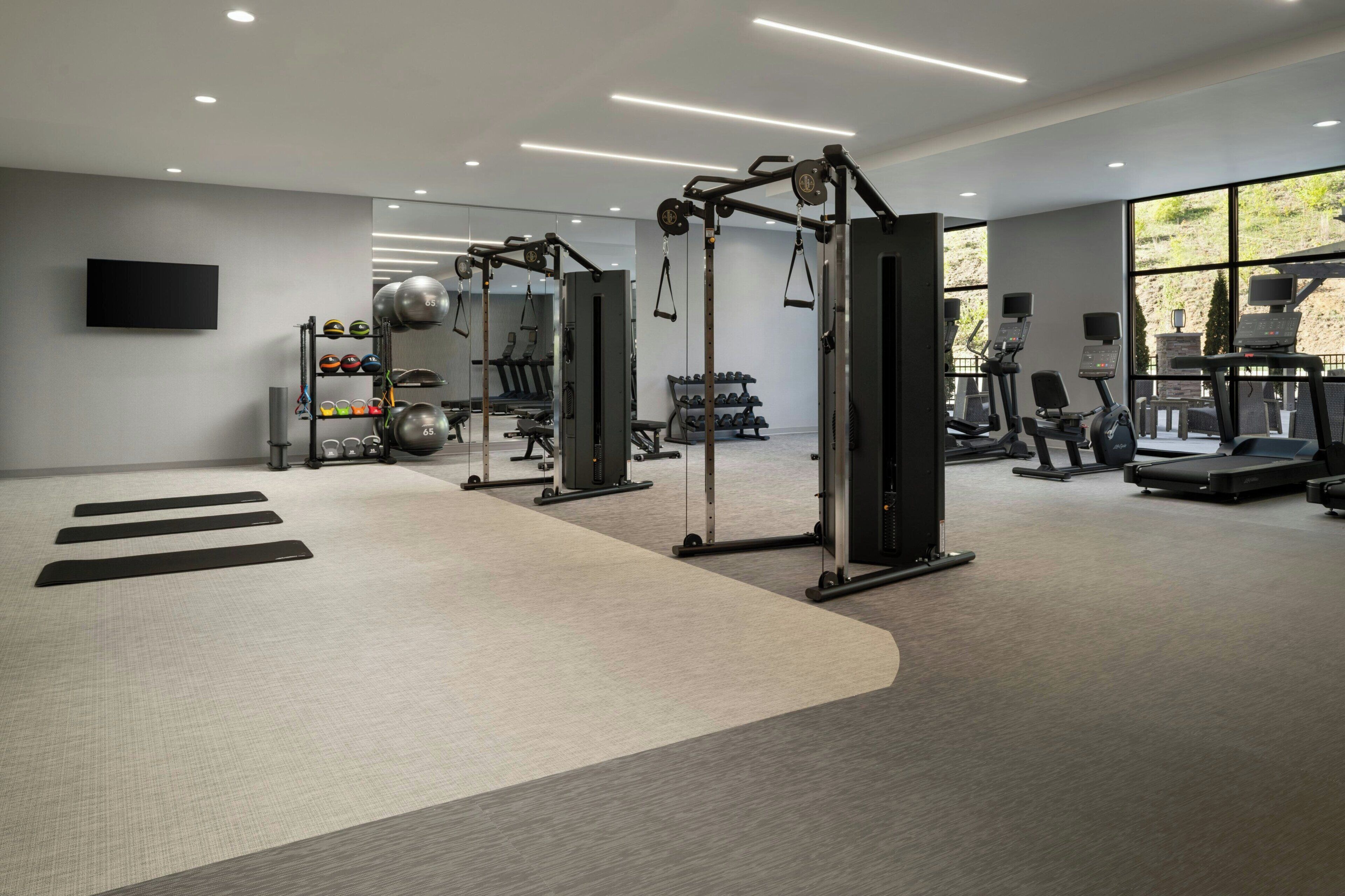 A modern gym with various exercise equipment, a wall-mounted TV, and a large window providing natural light.