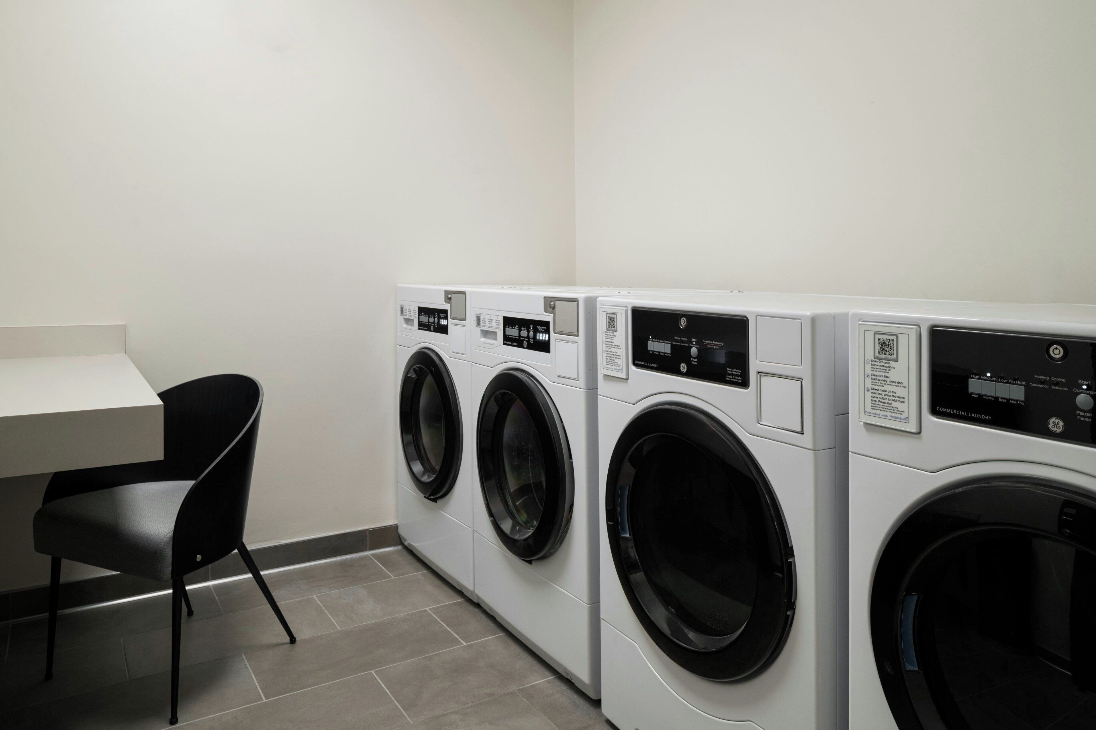 A laundry room with three front-loading washing machines, a black chair, and a white table.