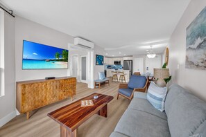 Ein modernes Wohnzimmer mit einem großen Flachbildfernseher, einem Holztisch, einem grauen Sofa und einer Küche im Hintergrund.