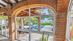 A covered patio area with brick arches and large windows overlooking a swimming pool and outdoor seating area. - Rancho San Jose - Barra De Santiago (Barra de Santiago)
