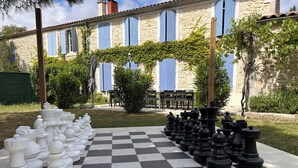 A large outdoor chessboard set up in a courtyard with stone buildings and blue shutters in the background.