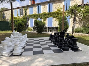 Un grand jeu d’échecs en plein air, installé dans une cour bordée de bâtiments en pierre et dont l’arrière-plan est constitué de volets bleus.