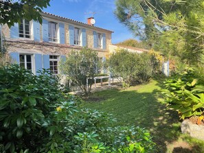 Un bâtiment en pierre avec des volets bleus, un jardin où poussent différentes espèces de plantes et un ciel dégagé.