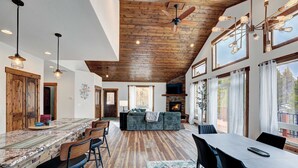 A spacious living area with a wooden ceiling, large windows, a fireplace, and a bar area. - Platte River Valley Cabin (Fairplay)