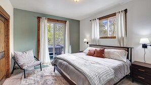 A bedroom with a bed, a chair, a nightstand, and a window with curtains. - Platte River Valley Cabin (Fairplay)