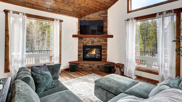 A cozy living room with a stone fireplace, wooden ceiling, and large windows. - Platte River Valley Cabin (Fairplay)