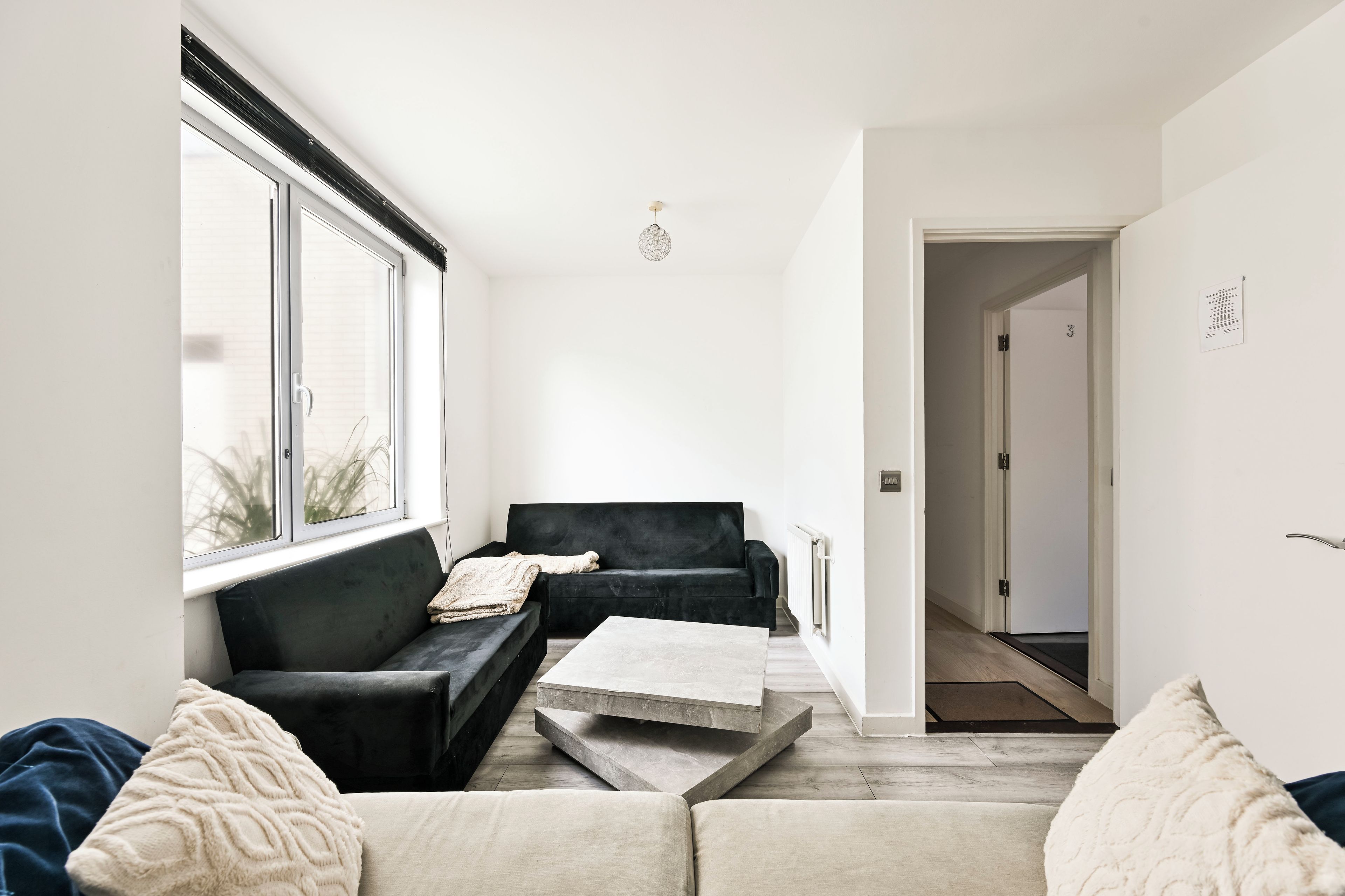 Een moderne woonkamer met een zwarte bank, een beige bank en een salontafel. Links is een raam en aan het einde van de kamer een deur.