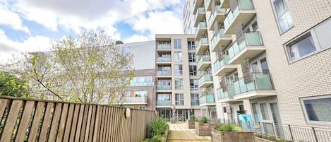 A modern apartment building with multiple balconies, a wooden fence, and a paved pathway.