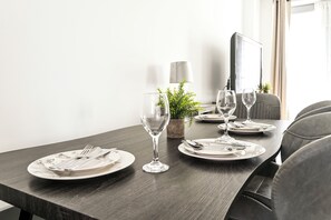 Una mesa de comedor dispuesta con platos, cubiertos y copas de vino, con una planta en maceta y una lámpara al fondo.