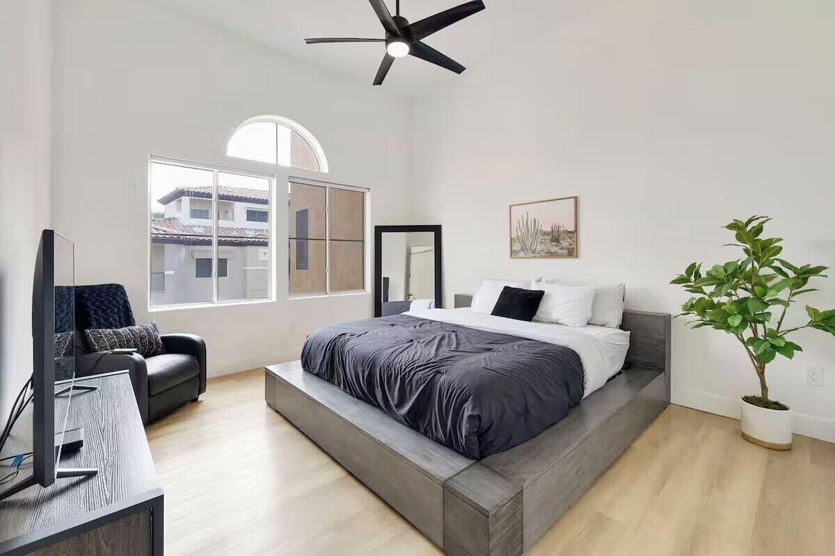 Quarto moderno com cama grande, escrivaninha, cadeira, ventilador de teto, janela e planta em vaso.