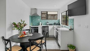 Uma cozinha compacta com armários brancos, painel de fundo azul-esverdeado e um espaço de refeições com mesa e cadeiras redondas.