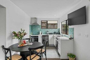 A compact kitchen with a dining area, a TV mounted on the wall, and a window with blinds. - Pataka Lookout (Taupo)