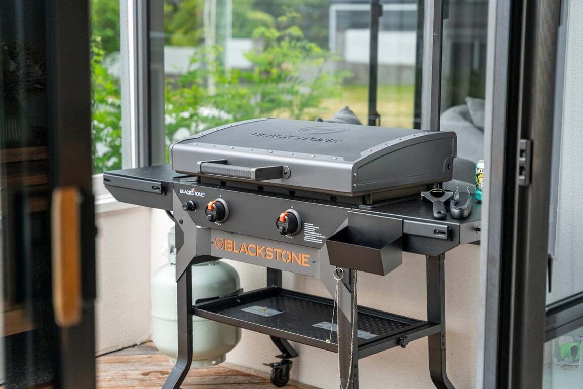 Una parrilla portátil BLACKSTONE con pantalla digital, sobre un carrito metálico cerca de una puerta corrediza de vidrio.