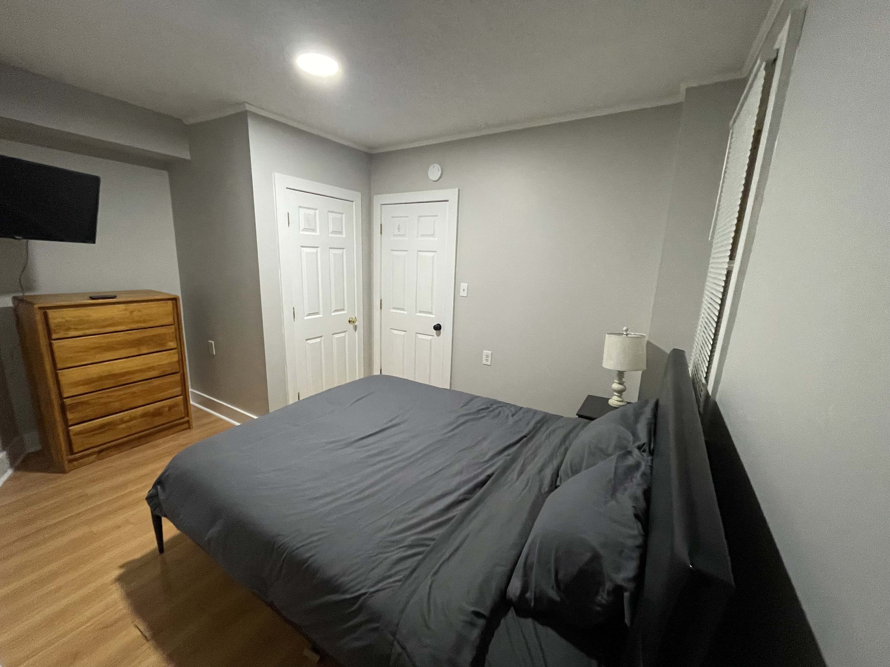 Un dormitorio con cama, cómoda, mesita de noche con lámpara y dos puertas.