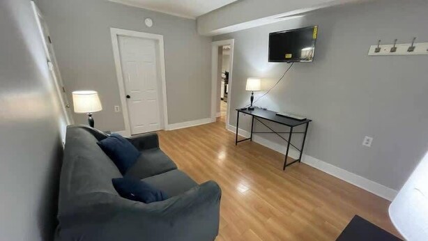 A living room with a grey sofa, a small table with a lamp, a TV mounted on the wall, and a wooden floor.