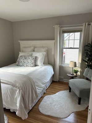 A bedroom with a bed, a chair, a small table with a lamp, and a window with curtains. - 3-bedroom house in a peaceful Saint Paul neighborhood with a fenced in back yard (Saint Paul)