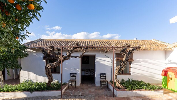 Un edificio con tetto in tegole, un ingresso coperto con sedie e un'area giardino con piante e un albero.