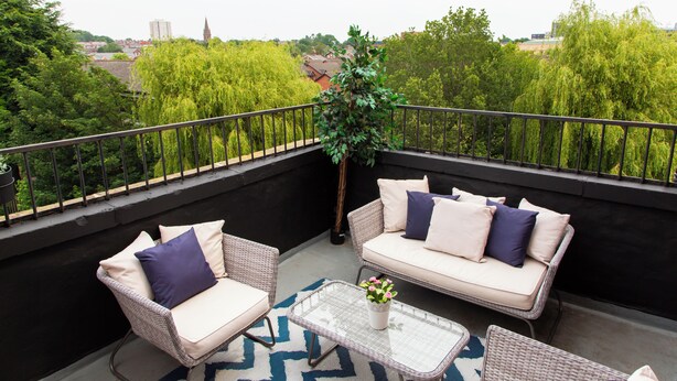 A balcony with wicker furniture, a glass table, and a view of greenery and buildings. - Residency apartments liverpool (Bootle)