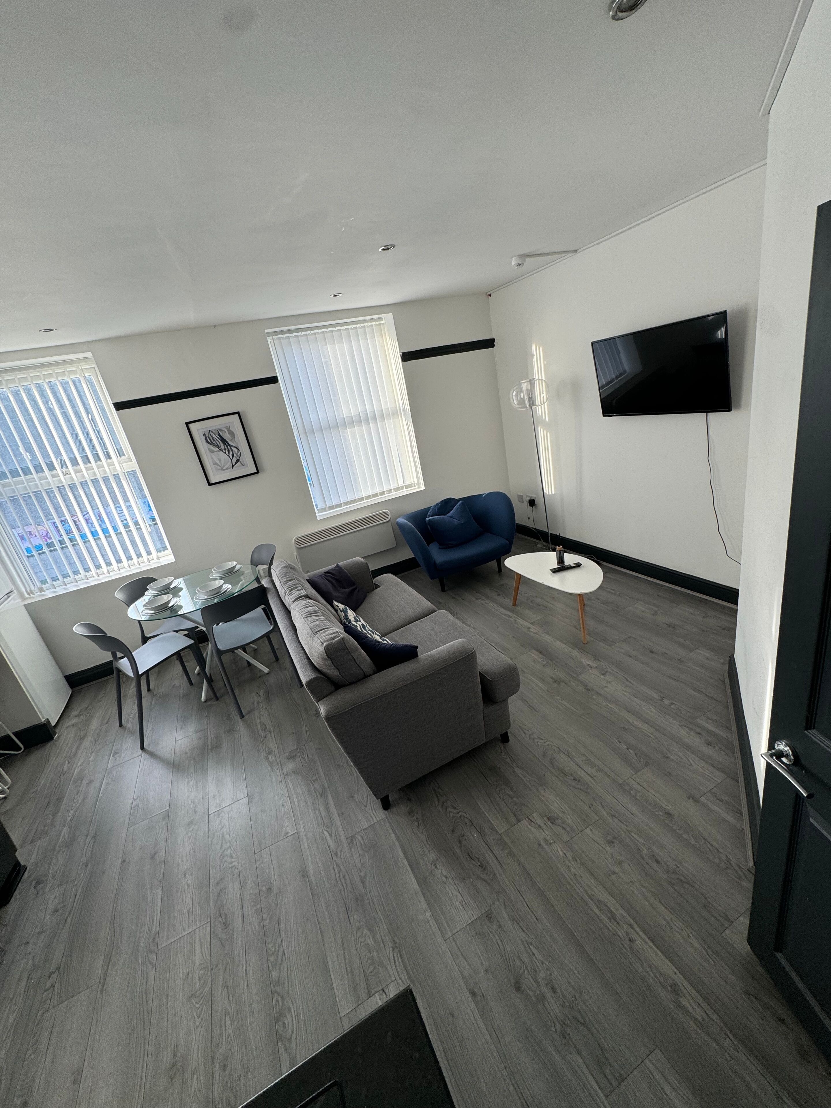 A modern living room with a grey sofa, a dining table with chairs, and a flat-screen TV mounted on the wall.