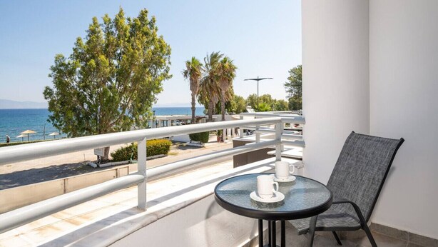 Een balkon met een glazen tafel en stoel, met uitzicht op een strand met palmbomen en een heldere lucht.