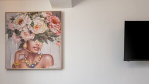 Een moderne kamer met een grijze bank, een flatscreen televisie aan de muur en een schilderij van bloemen en sieraden erboven.
