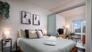 Une chambre moderne avec un grand lit, une table de chevet avec une lampe, une télévision et une vue sur la ville grâce à une grande fenêtre.