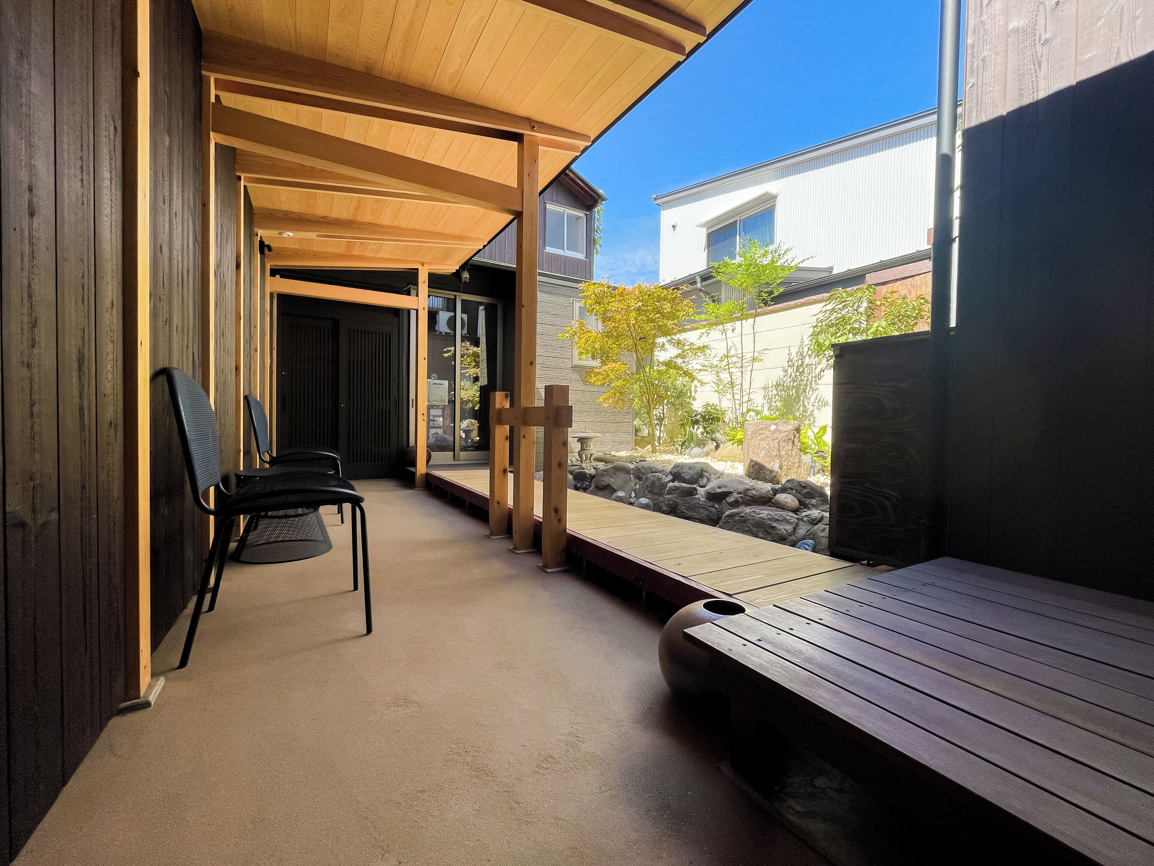 Un espacio de descanso al aire libre con techo, banco y sillas de madera, un sendero de piedra y vista a un edificio residencial.