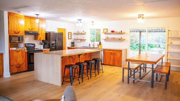 Une cuisine moderne avec des meubles en bois, un îlot central doté de tabourets, un coin repas avec des bancs et une vue sur la verdure par les fenêtres.