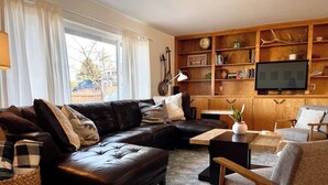 Un salon avec un canapé en cuir, une table basse en bois, un fauteuil gris et une bibliothèque.