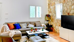 Un salon avec un canapé, une table basse et un téléviseur. On peut voir des plantes en pot sur la table basse et un mur en pierre au fond.
