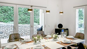 Un espace repas avec une longue table dressée pour quatre personnes, de grandes fenêtres offrant une vue sur la verdure et un éclairage moderne suspendu au plafond.