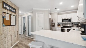 A modern kitchen with white cabinetry, a large island, stainless steel appliances, and a corkboard with a 'happy place' sign. - 30a Beach getaway, pool, Save 15% End of Summer (Santa Rosa Beach)
