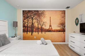A bedroom with a large wall mural of the Eiffel Tower, a bed with a white comforter, a bedside lamp, and a dresser with a television. - 9BR ChampionsGate Villa | Pool, Spa & Games (Four Corners)