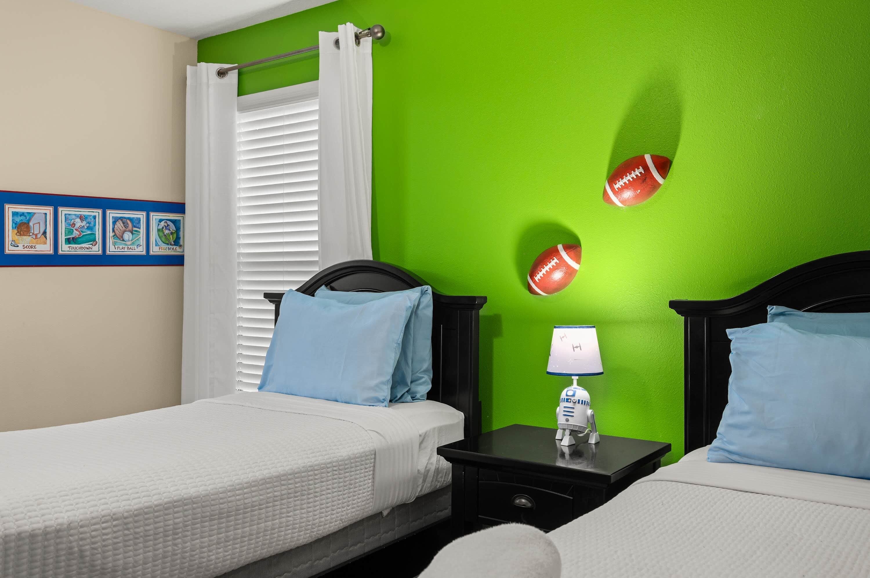 A hotel room with two beds, a nightstand with a lamp, and a green wall with sports-themed decorations.
