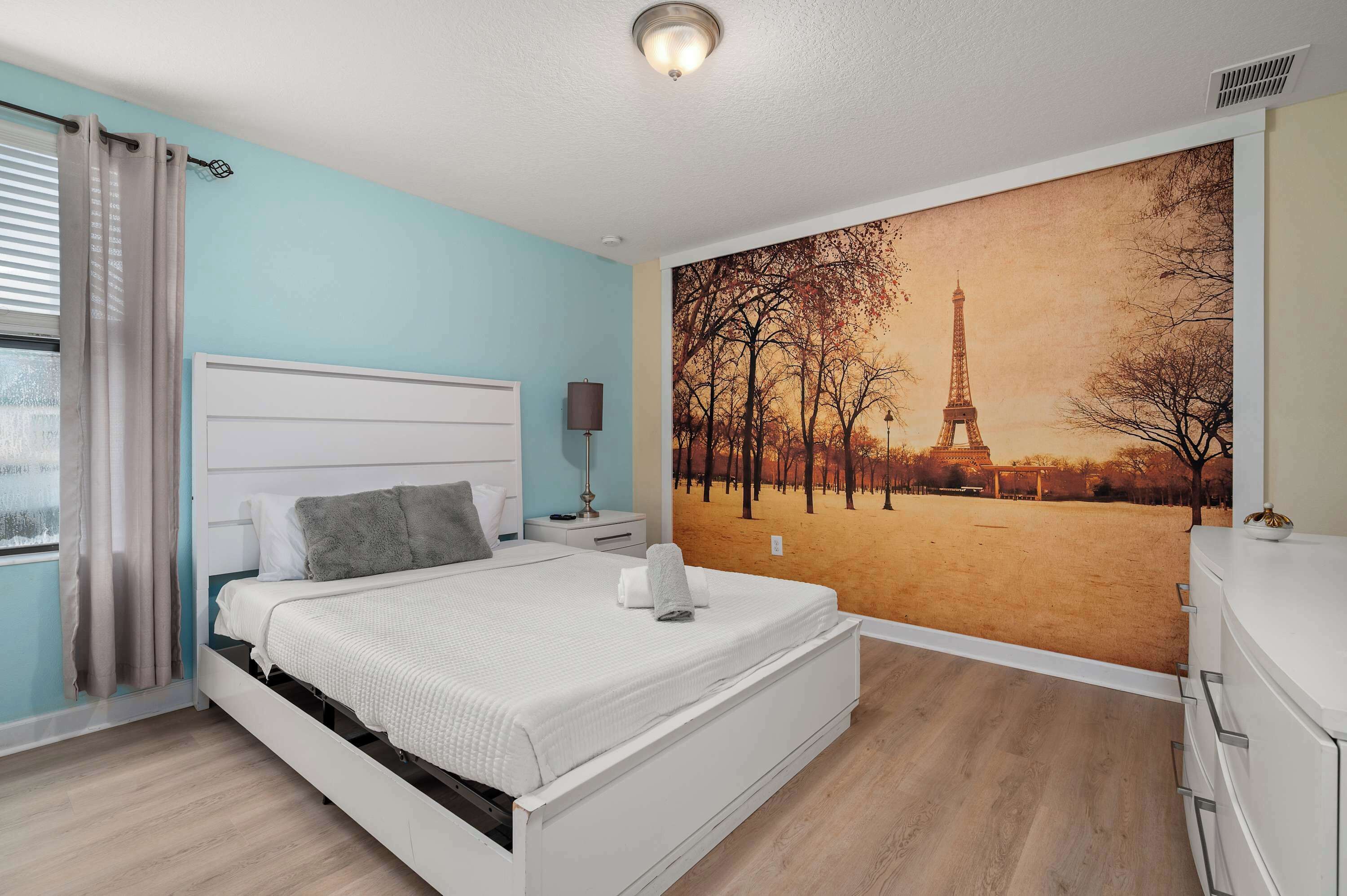 A bedroom with a bed, a nightstand, a lamp, and a wall mural of the Eiffel Tower.