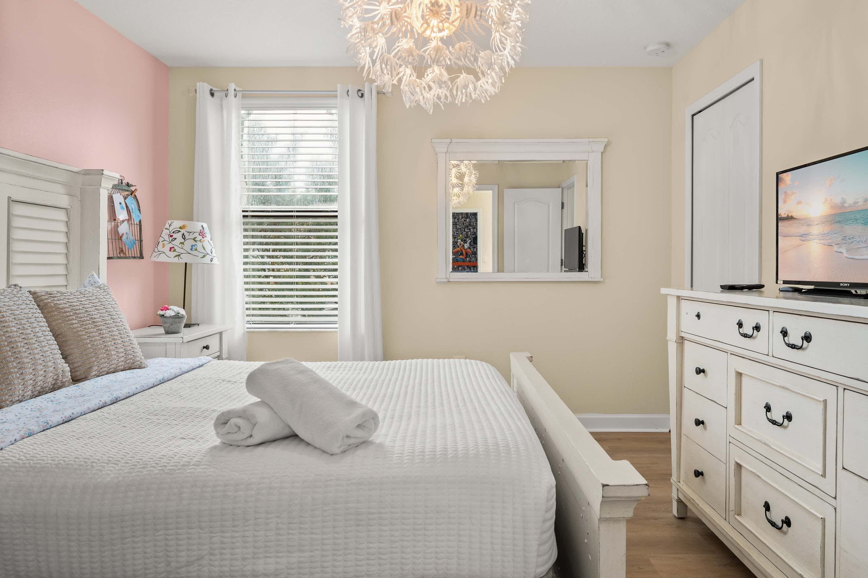 A bedroom with a bed, a dresser, a television, and a window with blinds.