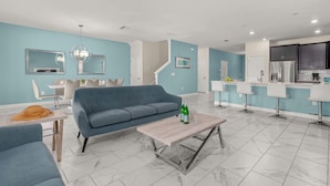 Una sala de estar moderna con un sofá azul, una mesa de centro con botellas y un comedor con sillas y una mesa.