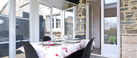 Un espace repas avec une table dressée pour quatre personnes, entouré de grandes fenêtres et de murs en pierre.