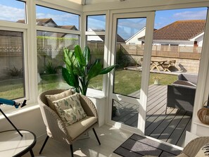 A sunlit room with large windows, wicker furniture, and a view of a backyard with a patio and outdoor seating. - Sunny Days: Bracklesham Bay Beach Bungalow – Sleeps 7, Parking & Dog Friendly (Bracklesham Bay)