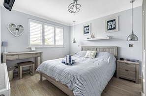 A bedroom with a bed, a desk, a chair, a bedside table, a nightstand, a lamp, and a window with blinds. - Sunny Days: Bracklesham Bay Beach Bungalow – Sleeps 7, Parking & Dog Friendly (Bracklesham Bay)